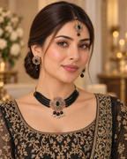 A woman wearing traditional Indian jewelry, including a black beaded choker necklace with a gold and green stone centerpiece, matching drop earrings, and a maang tikka, paired with a black and gold embroidered blouse.