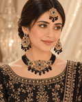 A woman wearing an ornate black and gold traditional jewelry set, including a necklace, matching earrings, and a headpiece, paired with a black embroidered top against a softly lit beige background.