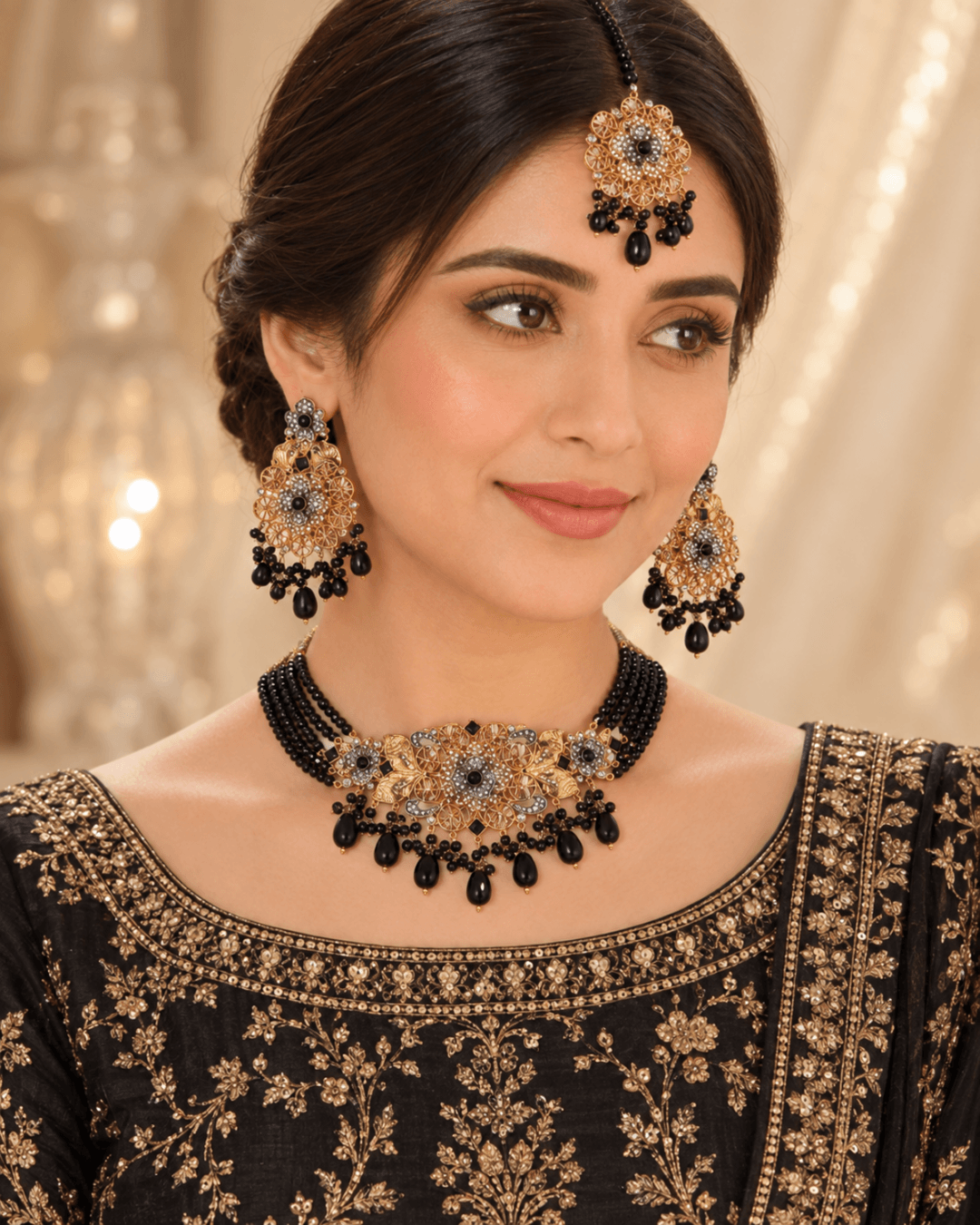 A woman wearing an ornate black and gold traditional jewelry set, including a necklace, matching earrings, and a headpiece, paired with a black embroidered top against a softly lit beige background.